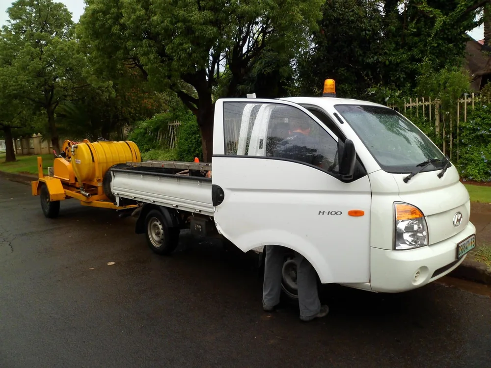 An ARPCO bakkie with a ladder on the deck towing a yellow high-pressure jetting trailer, a crew member at the driver's door